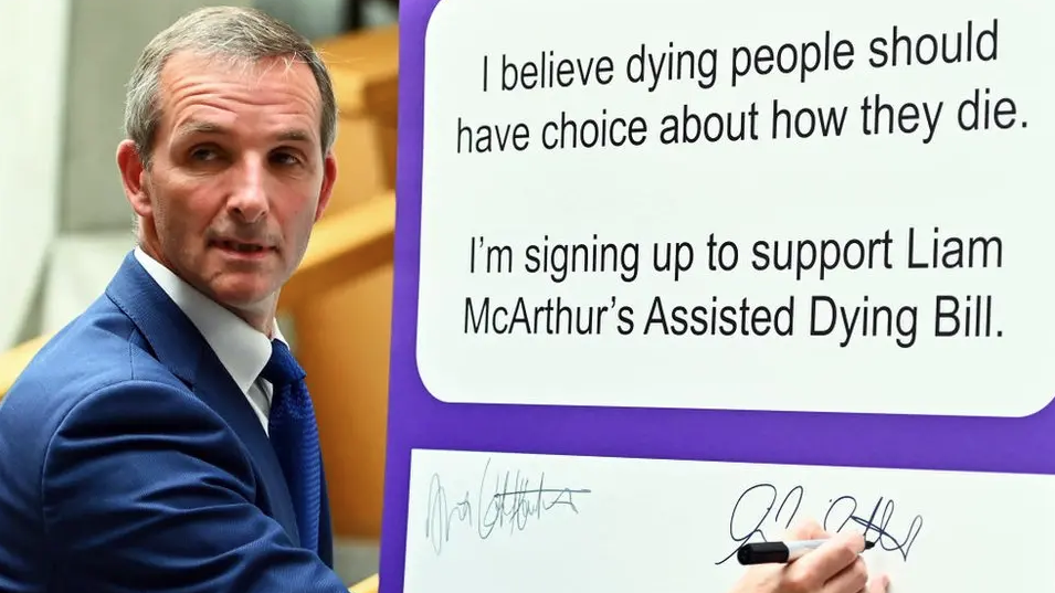 Liam McArthur, who has grey hair and is wearing a blue suit and tie and a white shirt, adds his signature to a list of MSPs who back his bill