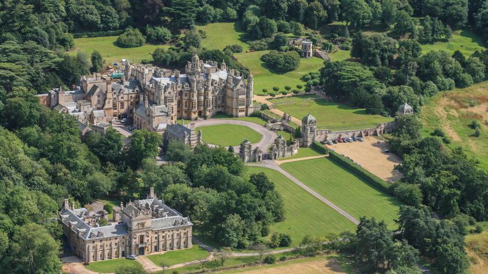 Aerial view of Harlaxton Manor, which was built in 1837. The house is set among trees and has a long driveway leading up to the entrance.