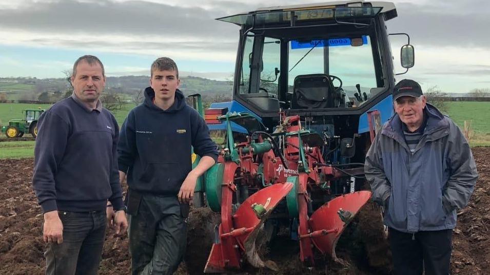 Three men stand in front of a blue tractor with plouging equipment attached. They are in a field, with grey skies and hills behind. There is another tractor in the distance.