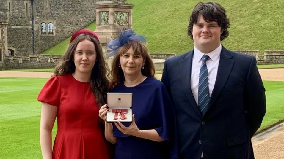 Fiona Devine, the founder of Alexander Devine Children's Hospice Service in the middle of her two children, wearing a navy dress and holding her MBE.