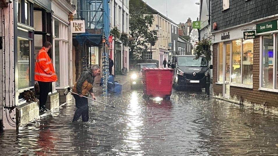 A view looking down the street. water can be seen almost knee deep as a person with a broom appears to be trying to clear a drain. A large red bin has been used to block traffic from coming through the road. 