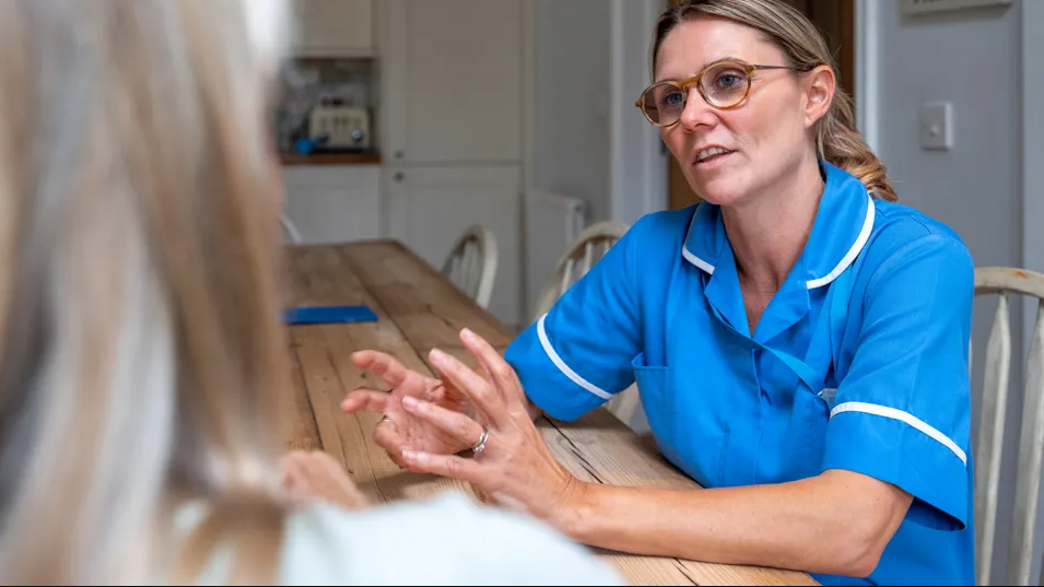 A nurse talking to a patient at home. We see the blonde hair of the patient and the front of the nurse who wears a blue uniform, has glasses and long hair swept back behind her ears. They sit at a wooden table