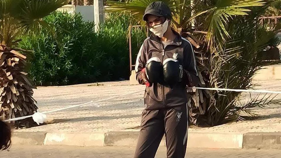 Malak Musleh standing up, wearing a black tracksuit, black boxing gloves, a black baseball cap and white facemask.