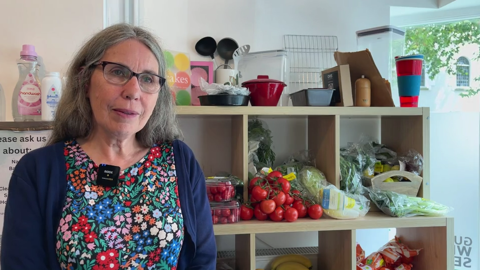 A woman with glasses and a flowery top with a navy cardigan. Behind her are shelves with fresh food.