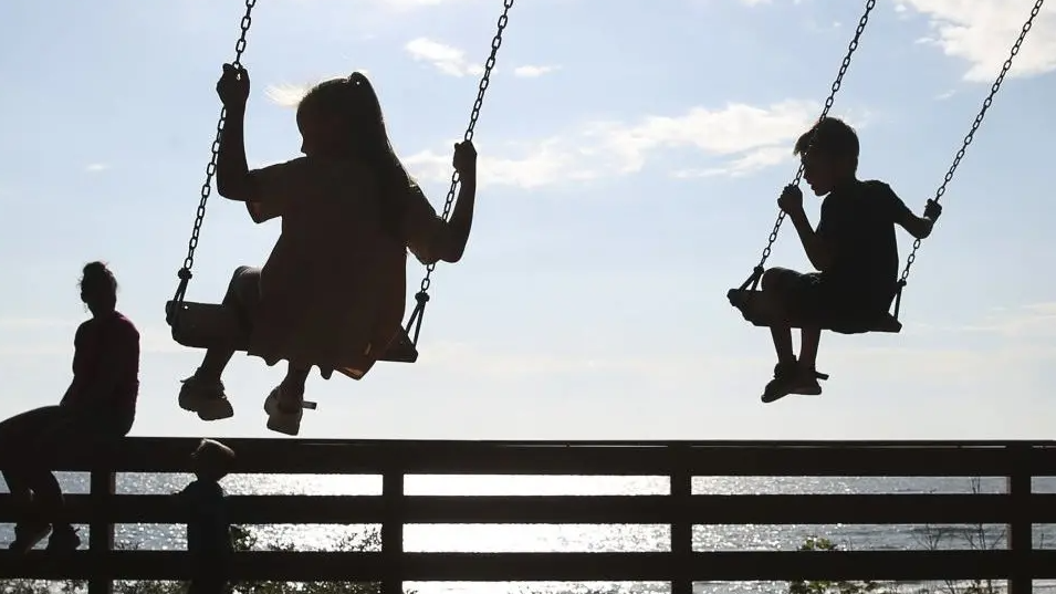 Children in silhouette sit on swings overlooking water with an adult on the left of the picture.