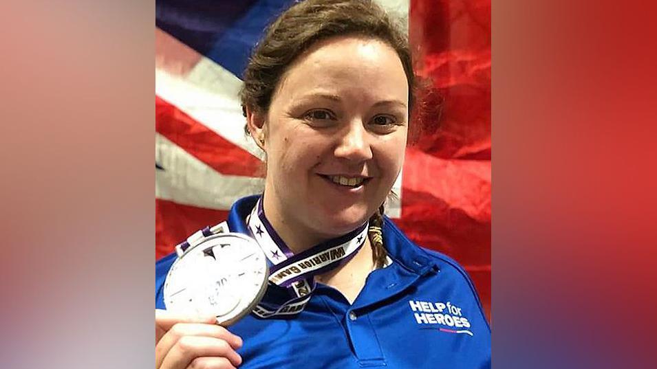 Gemma Kemble-Stephenson with brown hair in a plait wearing a Help the Heroes blue sports top smiling as she holds up a winning medal 