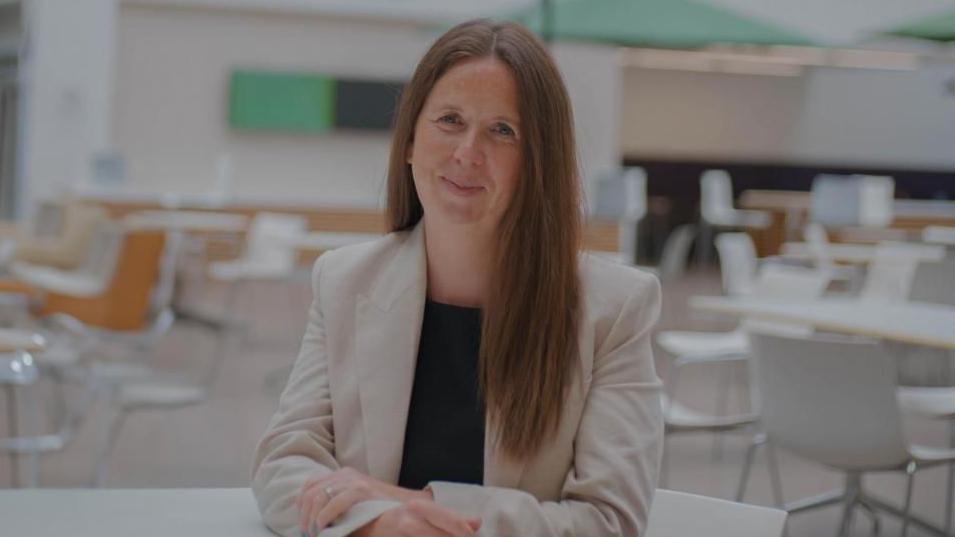 Lucy Townsend smiles at the camera. She has long dark hair, is wearing a beige jacket and sites in an empty cafe area