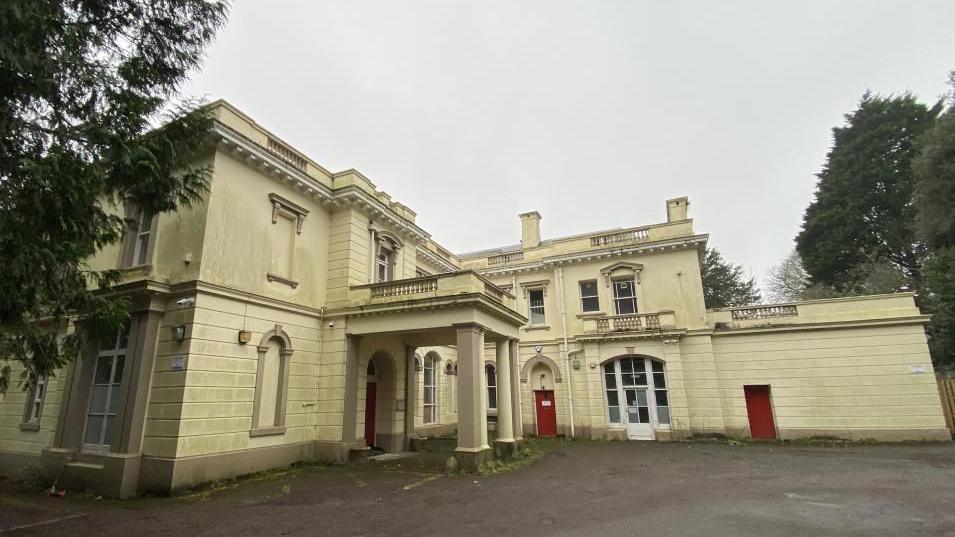 An outside shot of Pounds House located in Central Park in Plymouth. It is a large cream painted building with a canopy covering the front entrance with three red doors. It is surrounded by trees and has plenty of room for parking outside.