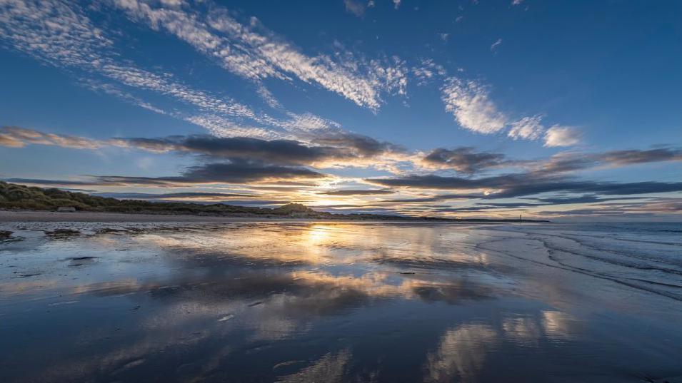 A shot of Lossiemouth West Beach at sunset. The sky is dark blue, with white grey and orange clouds in the distance. The sun can be seen going down far off in the middle of the shot. The sky is reflected by the water on the beach.