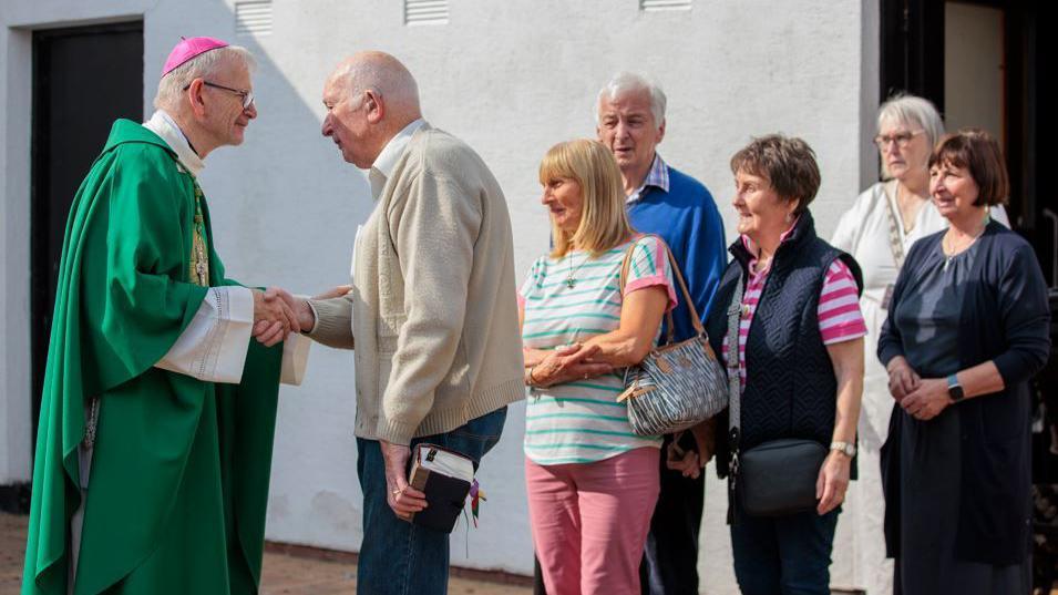 A bishop wearing a green cassock with a white neckline and he is wearing a purple skull cap. He has clear glasses and is greeting a line of six people coming out of Mass. At the front of a queue is a man with a cream cardigan  and grey hair who is holding the bishop's hand. Behind him a woman in pink jeans and a stripy green and white T-shirt has her arms folded. A man in a blue jumper and woman in a pink and white stripy T-shirt and dark gilet are behind them, followed by a woman with grey hair and glasses and wearing a white dress. The last person to the right is a woman with dark hair and a navy outfit.