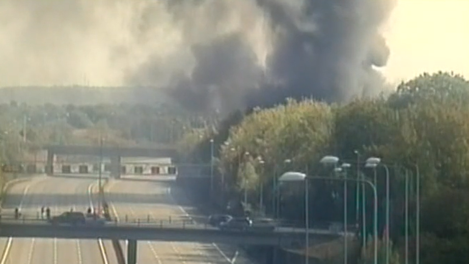 A large plume of smoke by the M25 motorway, with cars on the road across a bridge. Trees are to the left and all around the road. 