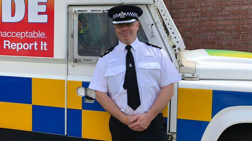 Ross Meredith, in his police uniform and cap, stands in front of a marked police van and smiles at the camera. 