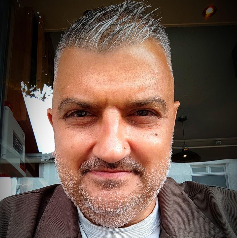 Picture of former policer officer Eurwyn Thomas. He smiles towards the camera in front of a glass window. He is of south-asian ethnicity, has brown eyes and a silver hair and beard. 