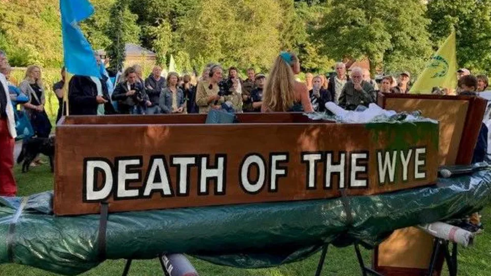 A coffin with Death of the Wye painted on it is lain on a surface outdoors with campaigners standing nearby on a sunny day by the river