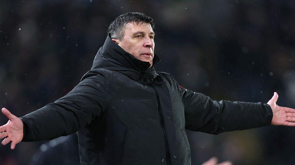 Hull City manager Sergej Jakirovic stands with arms outstretched in the rain, wearing black winter rain jacket