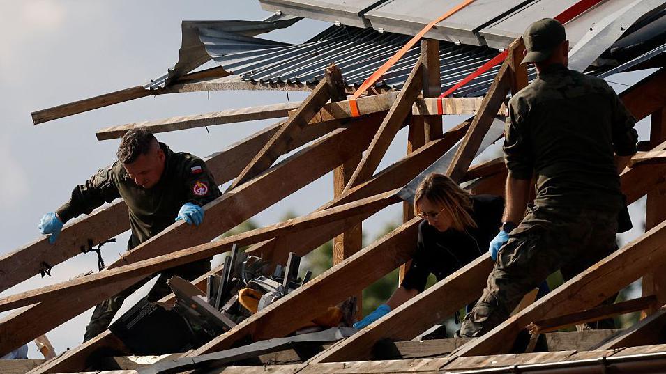 In Poland in September, three people inspect a damaged roof, where the wooden beams have been exposed. The roof was hit by falling parts of a drone that had been shot down.