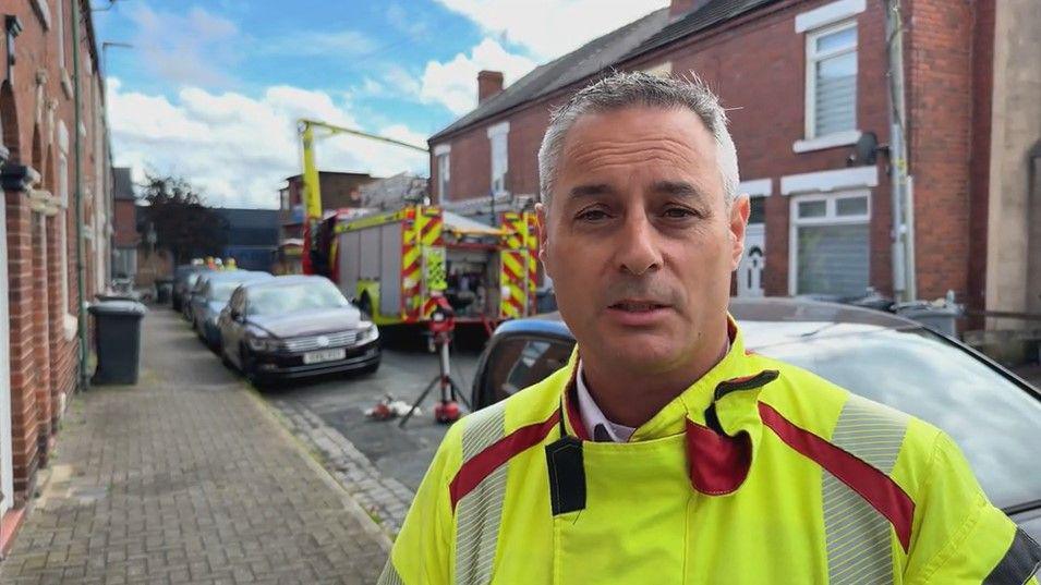 A fire officer wearing a hi-viz jacket and looking at the camera, with a fire engine behind him on a narrow terraced street