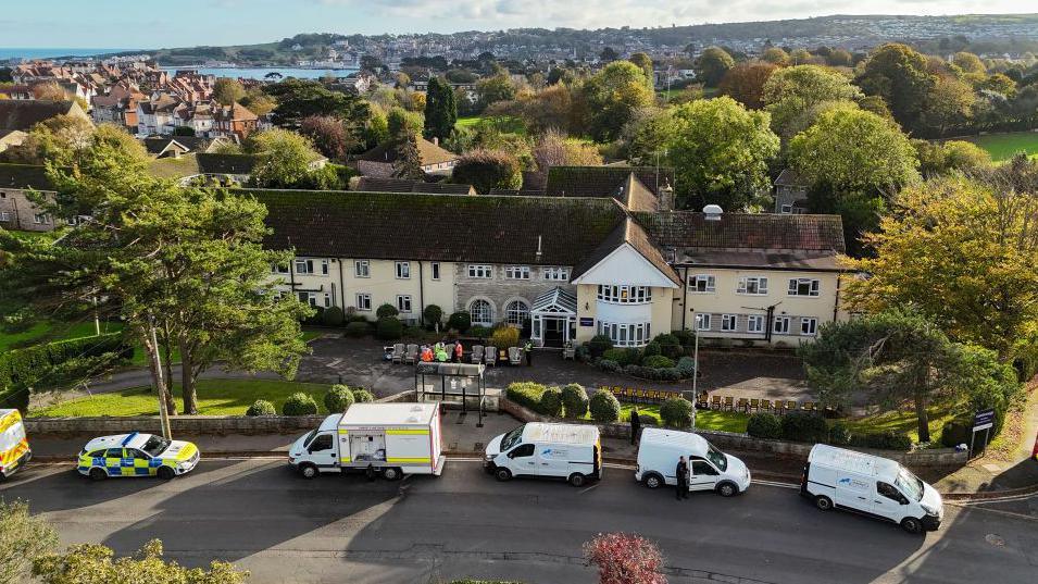 Drone image of the two-storey, cream-coloured care home with a line of police, fire and gas vehicles parked outside. There are trees behind the building and the town of Swanage in the distance beyond.