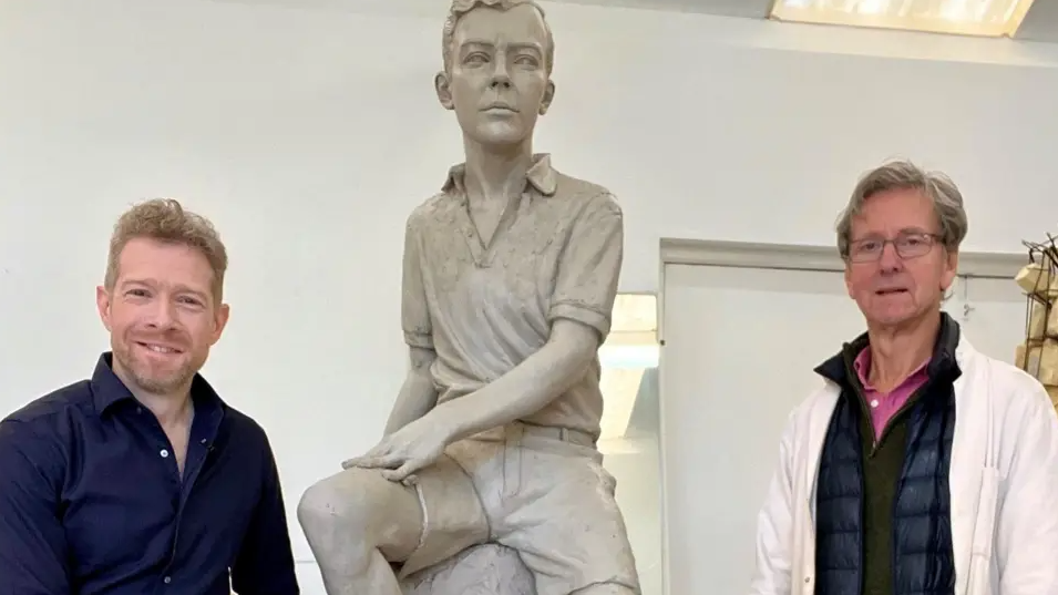 Zeb Soanes, wearing a blue shirt, and sculptor Ian Rank-Broadley, wearing a white jacket, stand either side of an alabaster sculpture of Benjamin Britten as a boy
