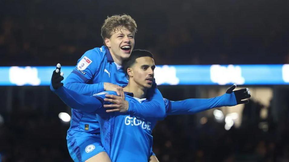 Two Peterborough United male players - one with his arms outstretched in celebration and the other jumping on his back.