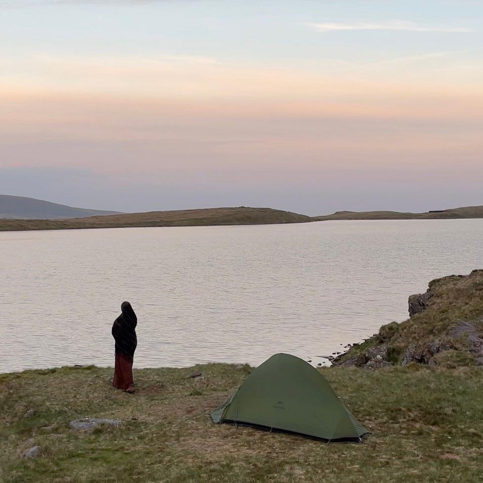 Amina stands by a vast river. Her back is facing the camera as she looks out to the water. There is a green tent set up to the right of her.