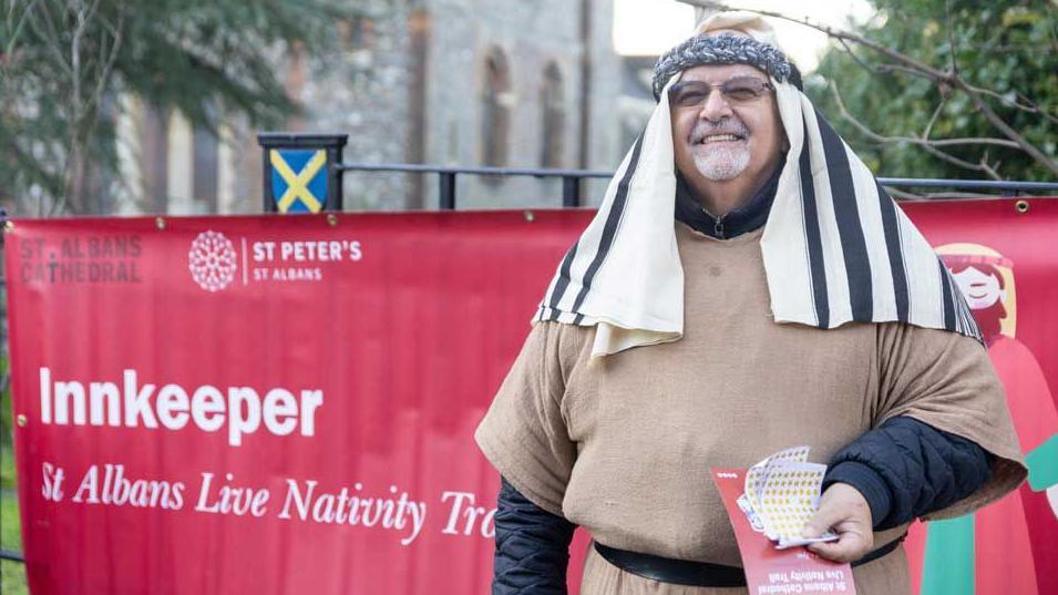 An actor playing the innkeeper stands in front of a red banner which says that he is the innkeeper. He wears a light brown tunic and a black-and-white headdress and is holding star stickers to give to the children