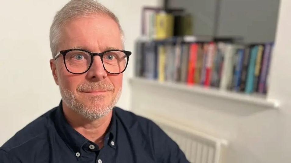 A man with grey hair and sporting a pair of black reading glasses. In the background there is a bookshelf with books placed on them.