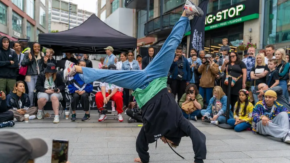 A crowd of people sit and stand on what looks like a public square. They are watching someone breakdance in the middle of the square. The dancer is currently on his hands with his legs splayed up in the air.