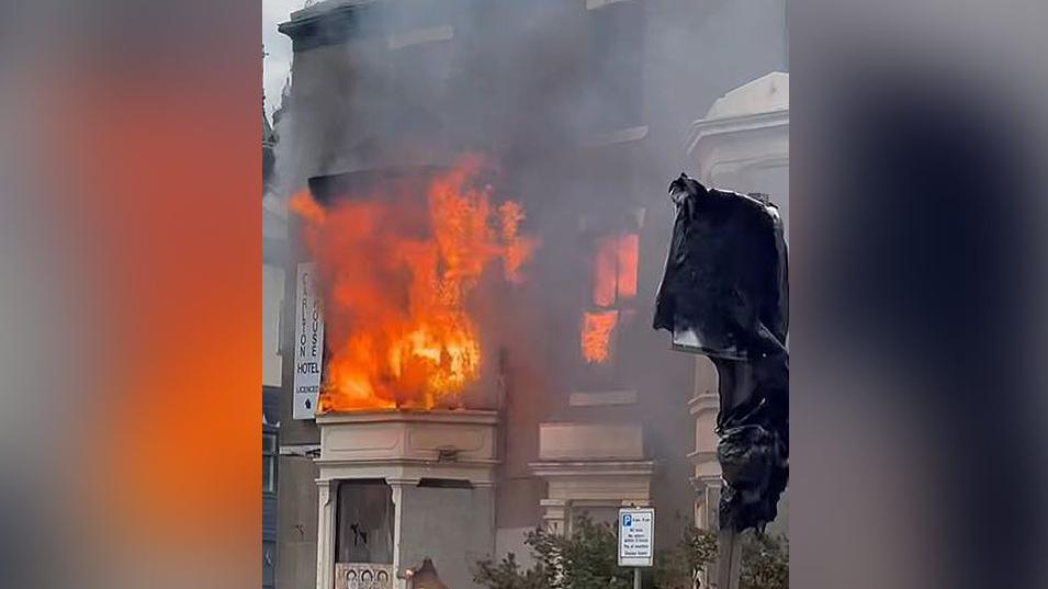 Fire burning in a former hotel in Blackpool. Bright orange flames and black smoke are billowing out of a first-floor bay window and second-first window.