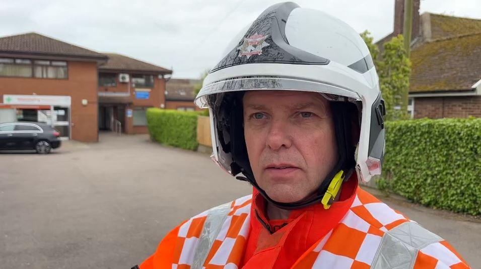 Chris Harding-Hook wears a fire officer's helmet and an orange hi-vis jacket. He is standing in front of a largely empty car park, with the medical centre behind.