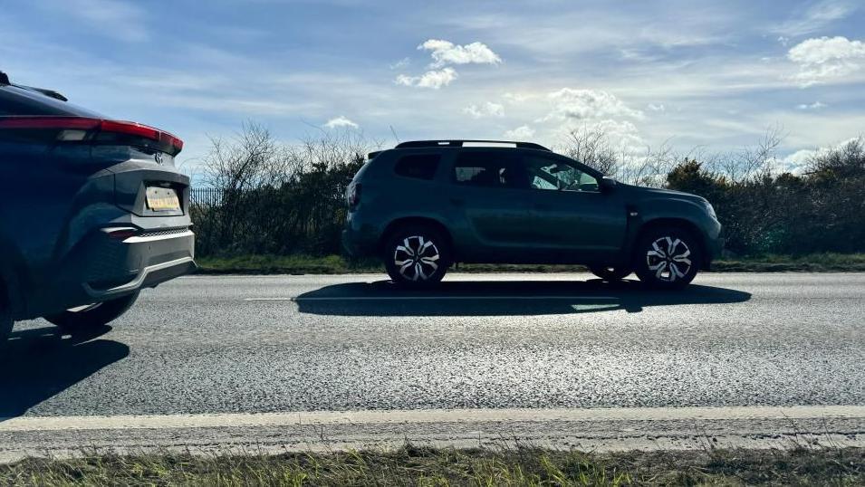 Two cars pass on the Acle Straight near Great Yarmouth. The picture shows the road surface and part of the verge. It also shows clouds in a blue sky and sunshine.