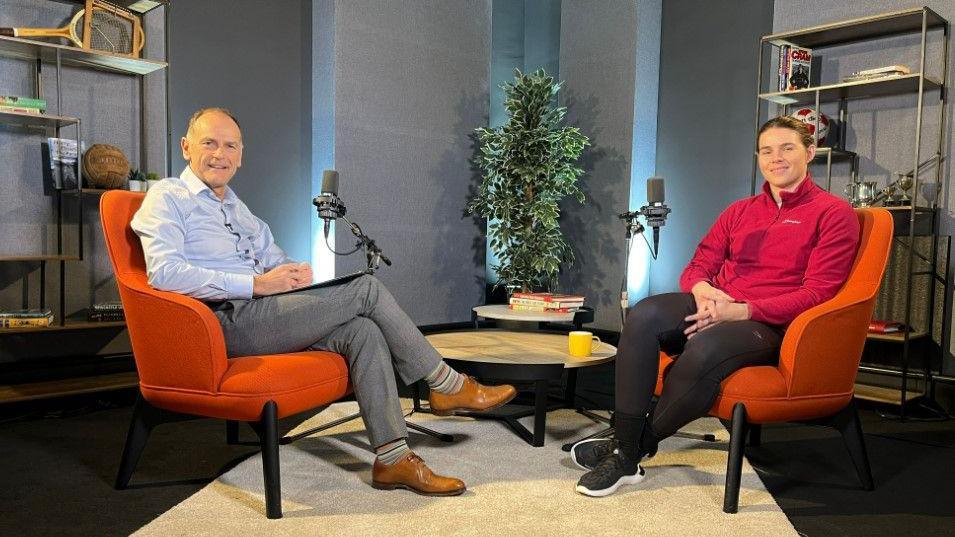 Jeff Brown and Savannah Marshall sitting on orange chairs and smiling in the podcast studio. Jeff is wearing a light blue shirt, grey trousers and brown shoes. Savannah's straight brown hair is tied in a bun. She is wearing black leggings, trainers and a red half-zip fleece. 