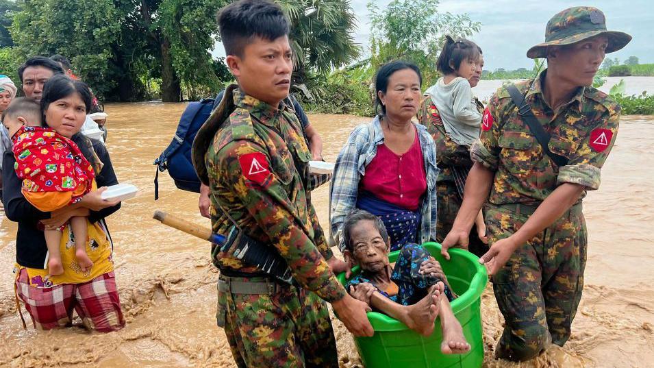 Two soldiers carry an elderly woman in a bucket as they wade through knee deep muddy water. Behind them several other civilians, carrying children, make their way through the flooding as well.