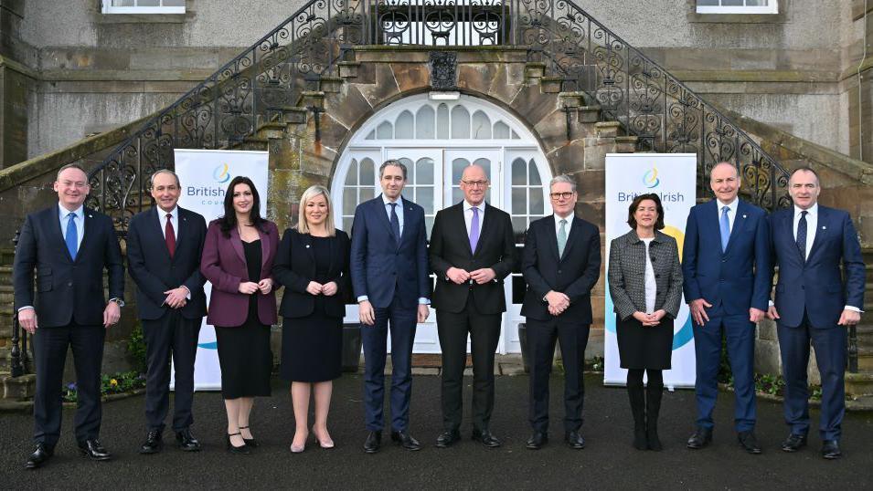 A family-style photograph of the 10 members of the British and Irish council standing outside a building with a white arched door. The seven men in the photo are all wearing dark suits, they include John Swinney who is bald with glasses and wearing a purple tie and Keir Starmer who has grey hair and is wearing a green tie. 