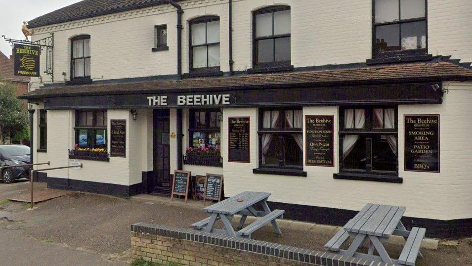 Exterior of The Beehive pub. The walls are painted cream and the window and door frames are painted black. Pub signage is on the walls and there are grey benches out the front.