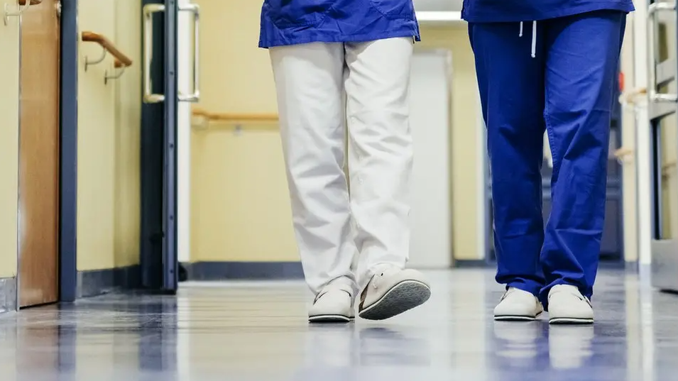 Two NHS staff walk along a corridor of a ward. Their faces are not visible, just see their legs.