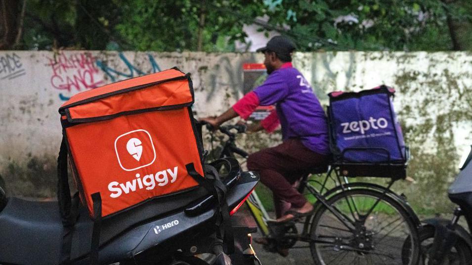 A Swiggy branded delivery box mounted on a motorcycle in Mumbai, India, behind which is a Zepto delivery man on a bicycle.
