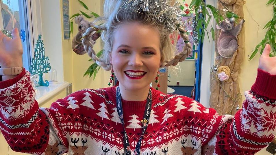 Hannah Gidman smiles at the camera with her hands up. She is dressed in a Christmas jumper with her hair in plaits and wrapped in tinsel. 