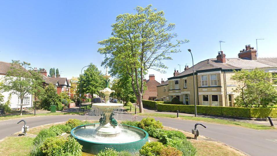 An elaborate fountain on a grassy roundabout is silhouetted in the early morning sunshine, behind it there is a tall tree and some large Victorian yellow brick houses. 