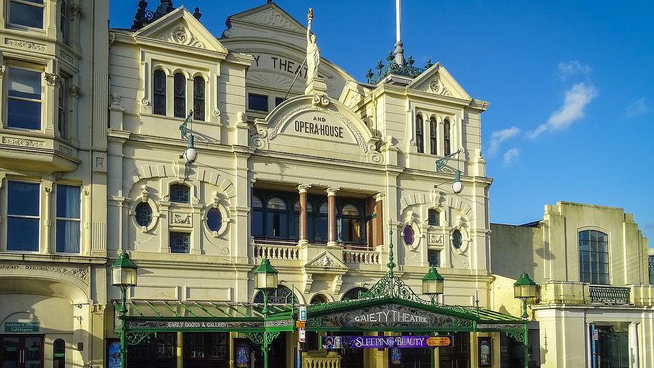 An ornate, Victorian-era theatre with a decorative, cream-coloured facade. "Gaiety Theatre and Opera House" is written on the front above the entrance, which is below a Greek statue holding a torch aloft. 