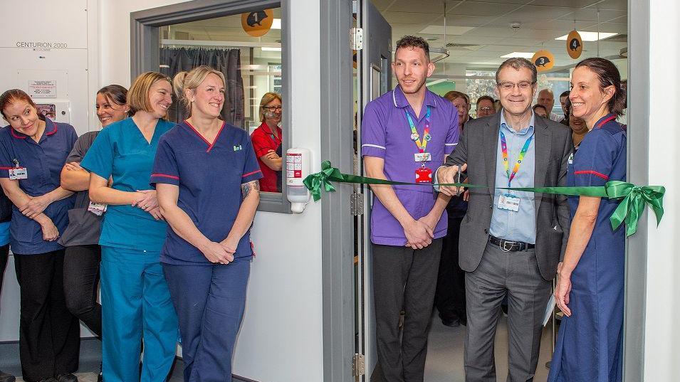 A group of people wearing scrubs smiling as a man in a suit cuts the ribbon at the entrance to a hospital ward.