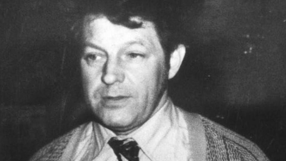 A black and white image of a man with dark hair. He is wearing a cardigan and a shirt and tie