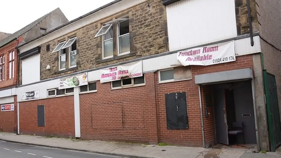 A street, showing a red brick building with stone on the second floor. Signs say function room available. Windows are open on the second floor. On the ground floor,  a door is wedged open.