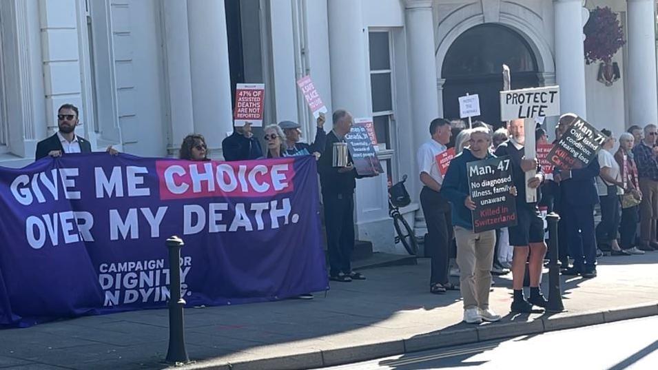 A large purple banner saying "give me choice over my death" is held up on the left. On the right people hold smaller signs, one reads "protect life".