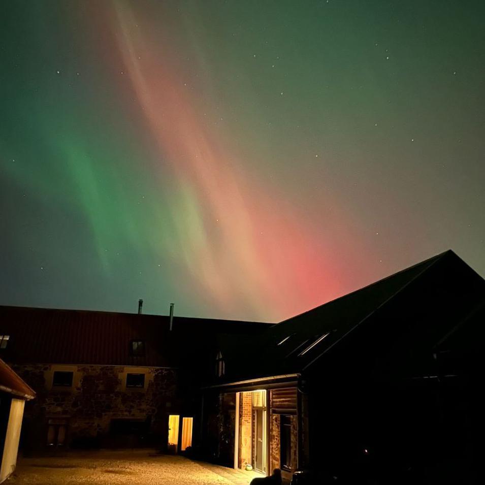 A house with lights on, with the aurora in colours of green and pink behind it.