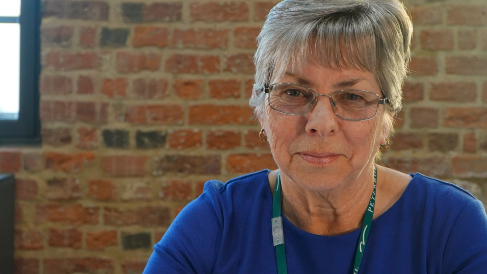 Geraldine stares at the camera. She wears a dark blue top and a green lanyard. Behind her is a brick wall with windows either side.