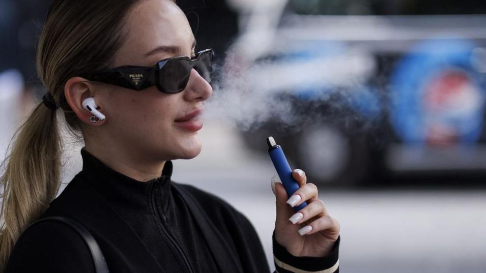 A young woman wearing a tracksuit top, Prada sunglasses and Airpods uses a blue-coloured disposable vape on a London street