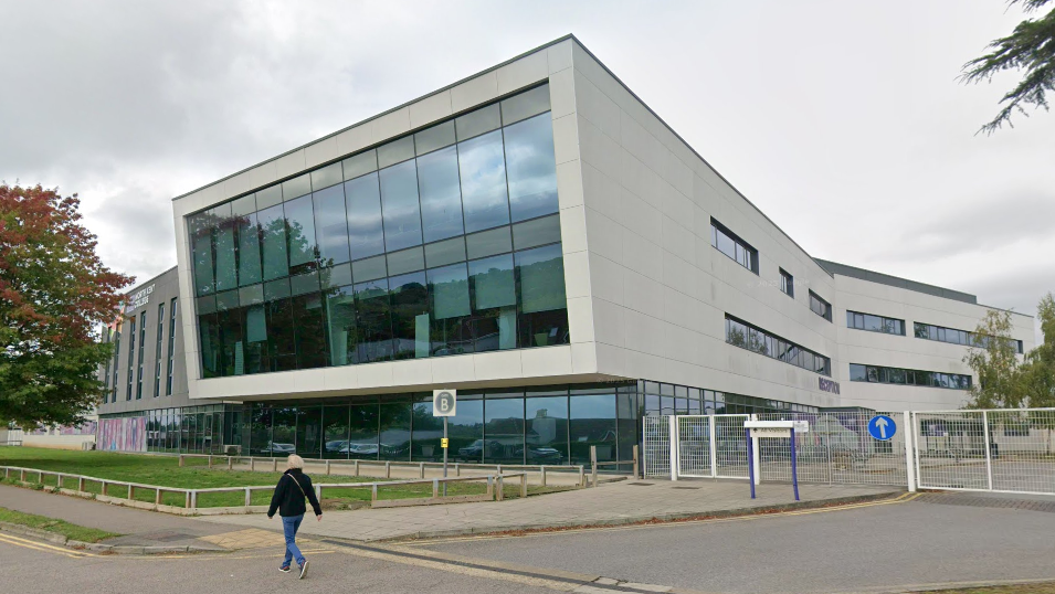 A college building with a large glass front and grey walls. A person is walking in front of the building.