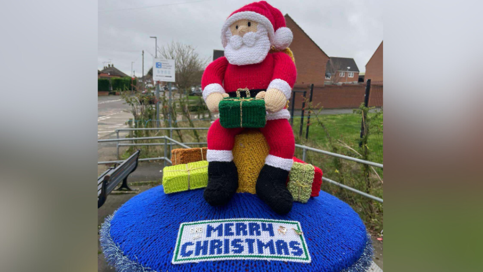 A knitted postbox topper depicting Santa Clause sitting on a chair surrounded my gifts with a blue base that says 'Merry Christmas' 