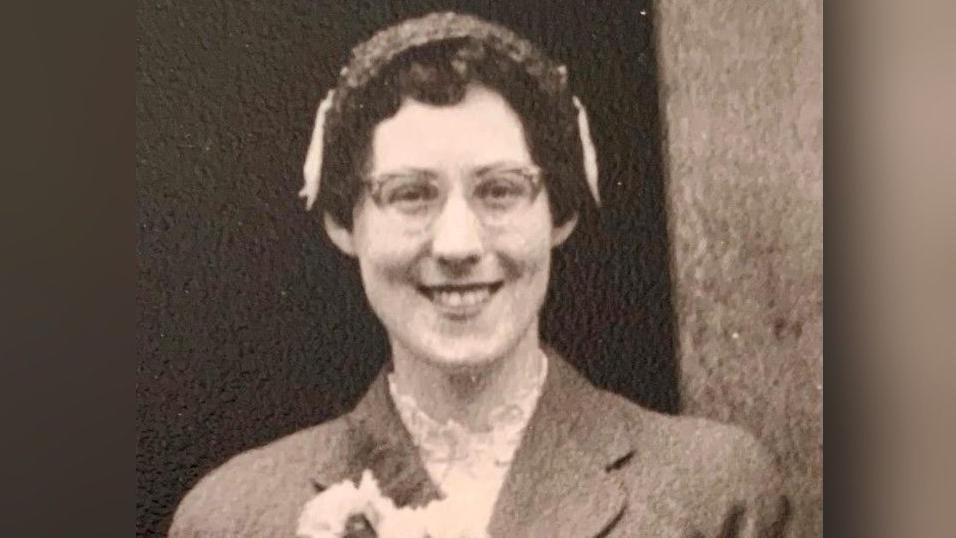 An old black and white photo of a woman with short black curly hair and glasses, who is smiling at the camera.